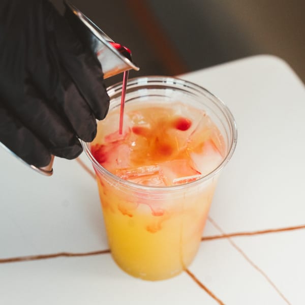 A gloved hand pours red syrup into a clear cup of iced orange drink.