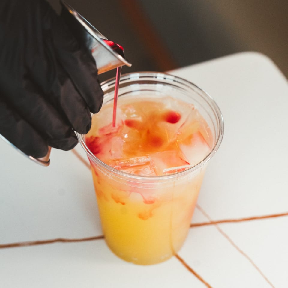 A gloved hand pours red syrup into a clear cup of iced orange drink.