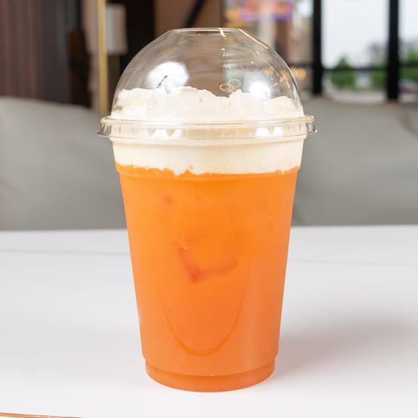 Orange drink with a creamy white foam topping in a clear plastic cup with a dome lid.