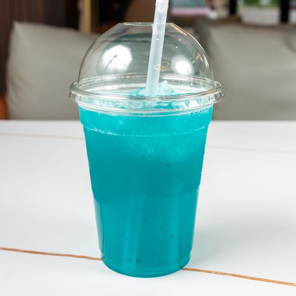 Bright blue slushy drink in a clear plastic cup with a dome lid and straw on a white table.