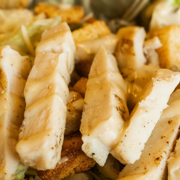 Close-up of grilled chicken strips and croutons with dressing, part of a Caesar salad.