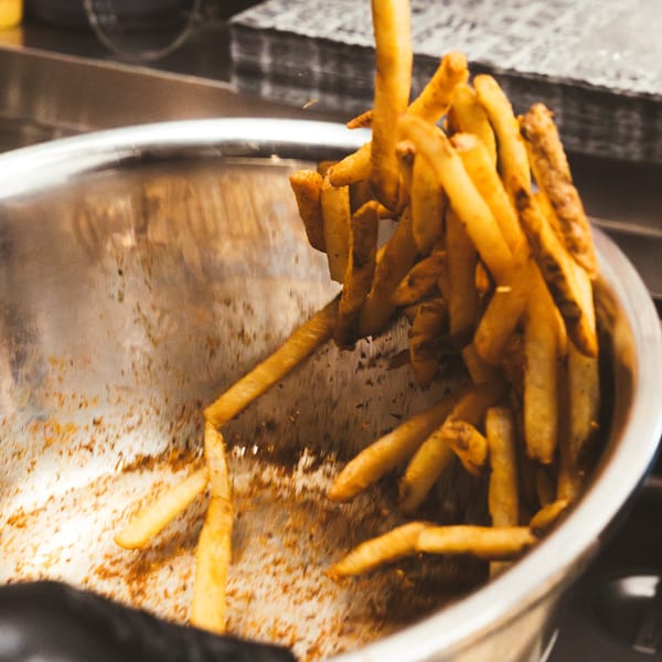 Golden-brown french fries being tossed in a large metal bowl, coated with seasoning.