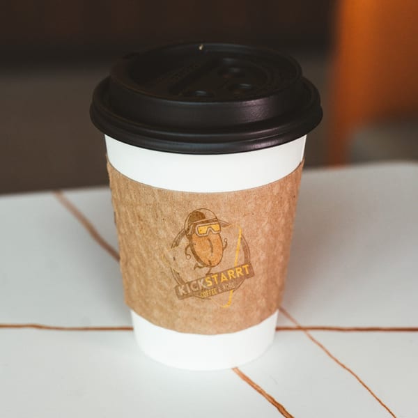 A white coffee cup with a brown sleeve featuring the "Kickstarrt Coffee & More" logo.
