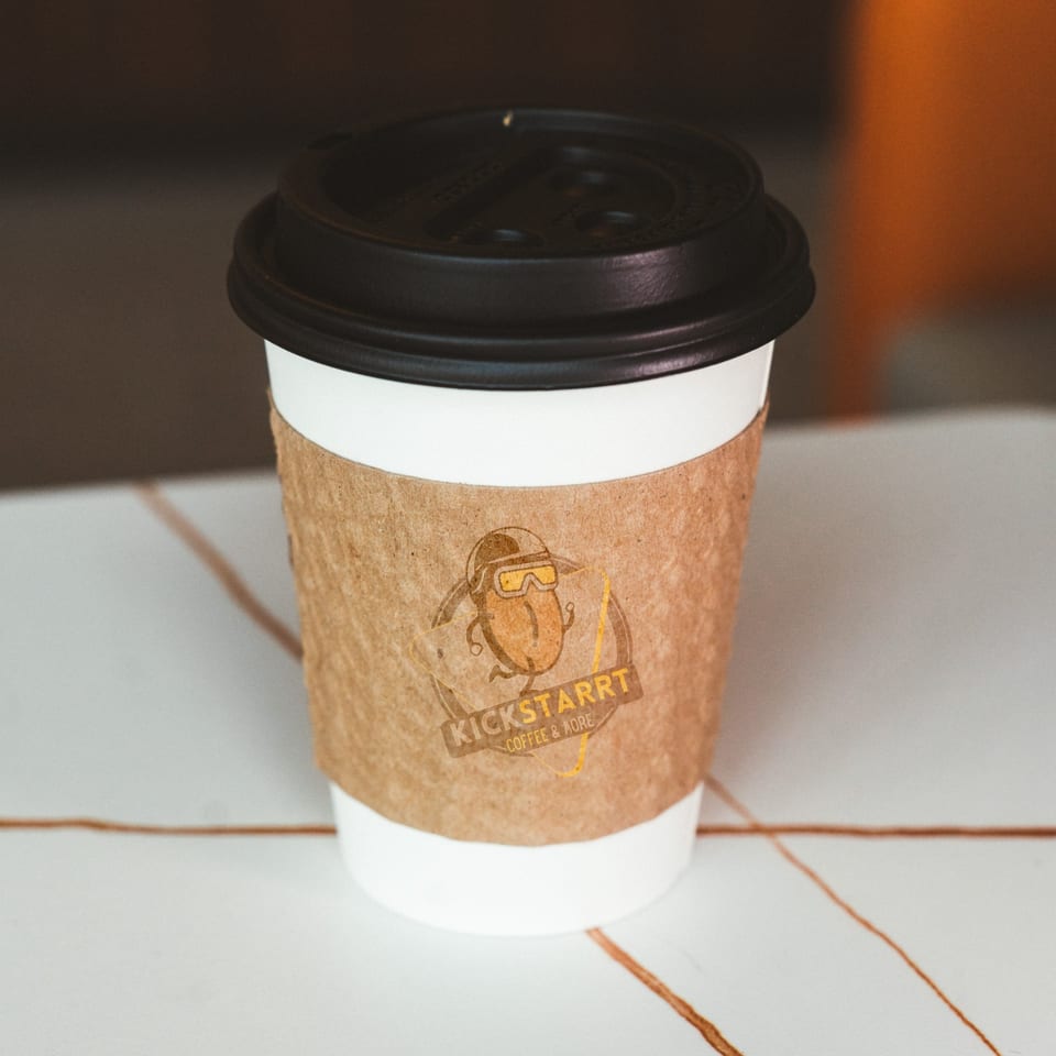 A white coffee cup with a brown sleeve featuring the "Kickstarrt Coffee & More" logo.