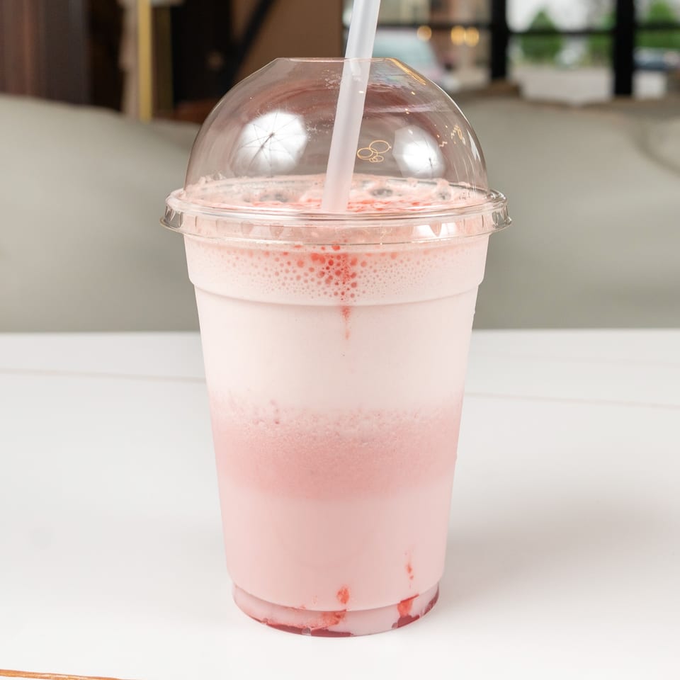 Pink strawberry milkshake in a clear plastic cup with a dome lid and straw on a white table.