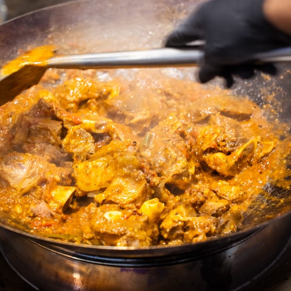 A chef stirring a large pot of simmering, golden-brown curry with meat and bones.