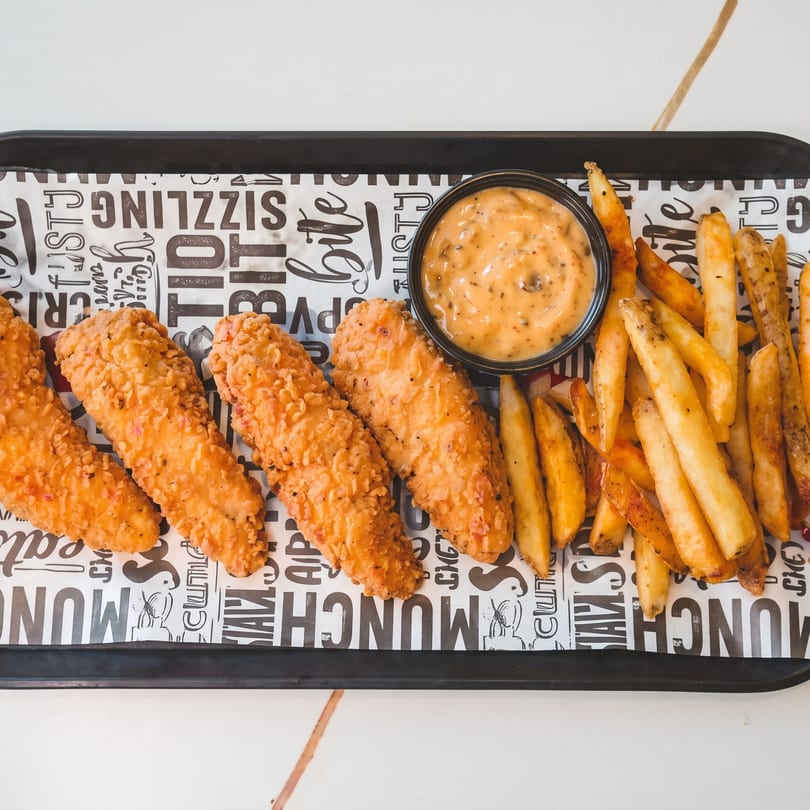 Crispy chicken tenders, golden french fries, and a dipping sauce on a black tray with patterned paper.
