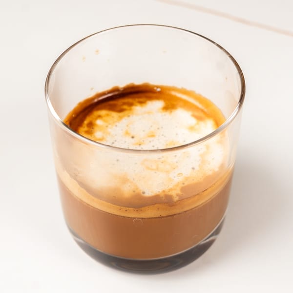 Close-up of a glass of espresso with a thick layer of crema on a white surface.